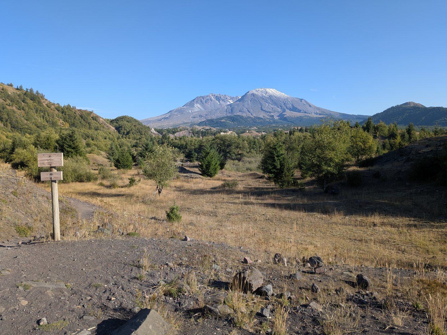 Mount St. Helens in the distance beyond a dusty trail and a wooden sign