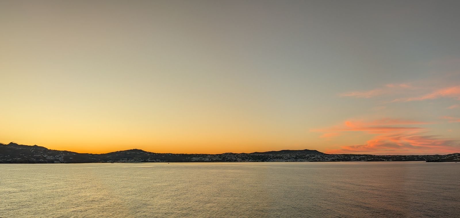 Sunset over calm water with a hilly coastline on the horizon